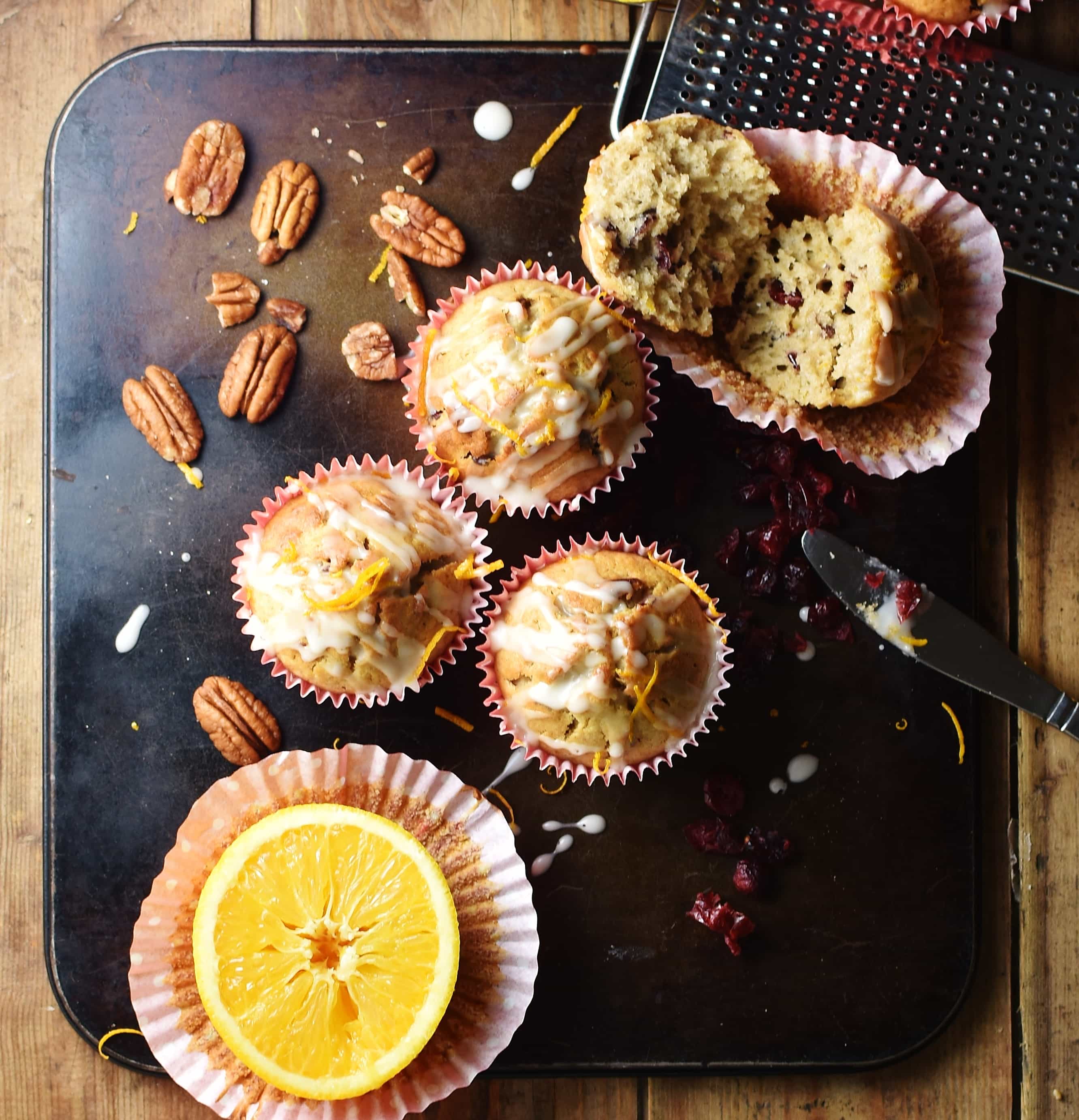 Muffins, dried cranberries, halved orange on top of paper case, pecans and fruit zester on top of dark tray.