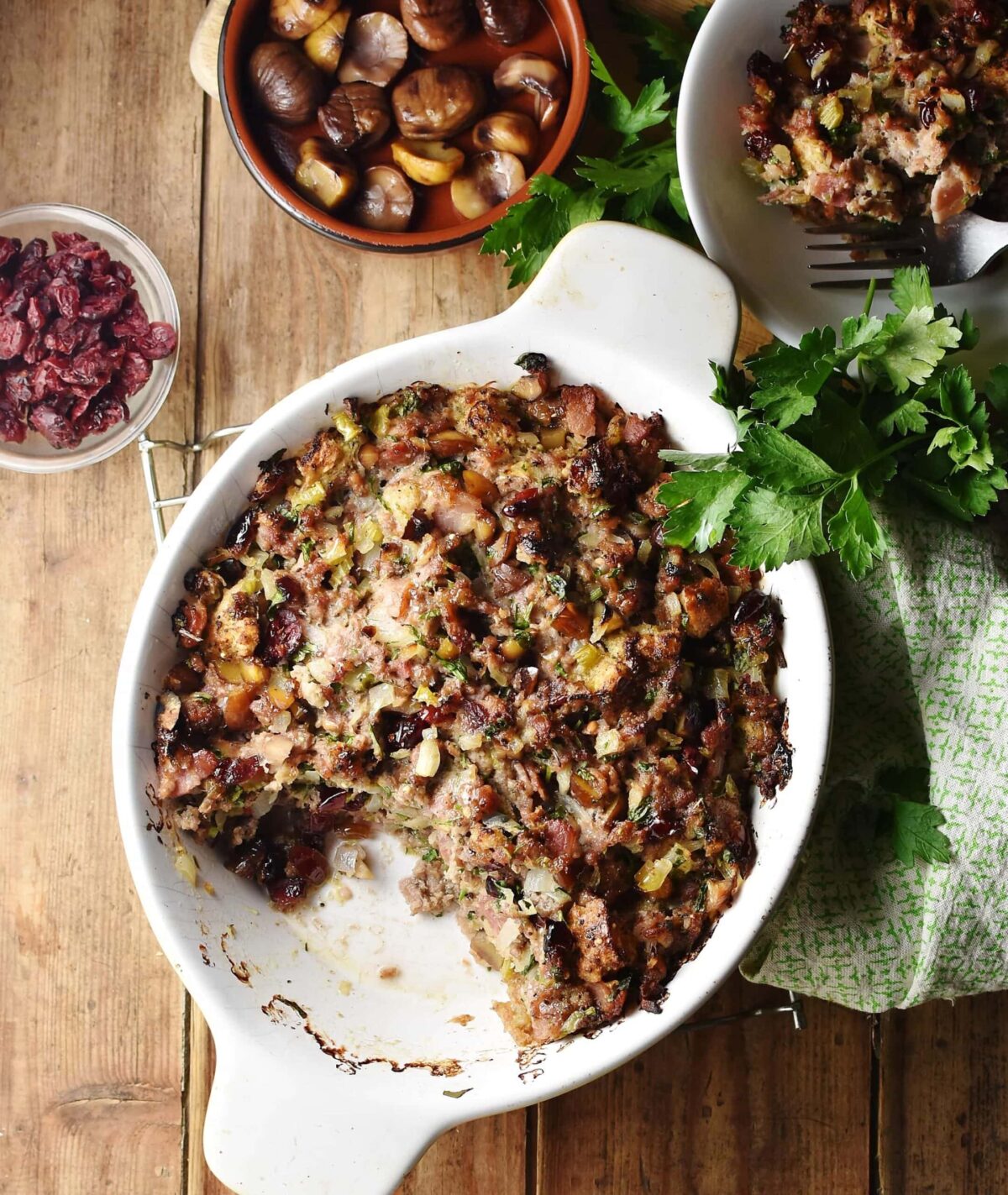 Top down view of turkey stuffing casserole in white oval dish, with green cloth to the right, casserole in white bowl, chestnuts in brown dish and dried cranberries in small dish in background.