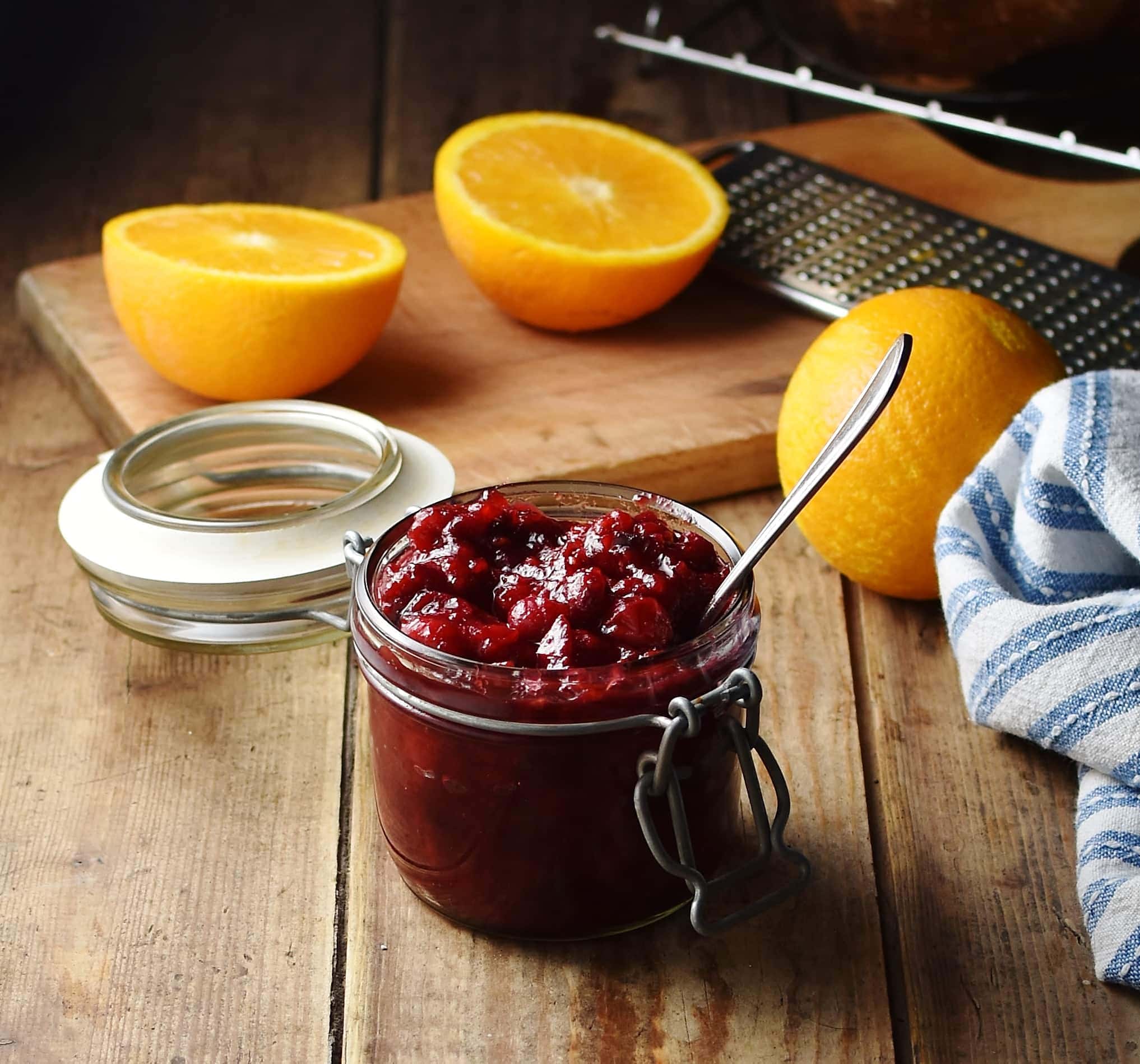 Side view of cranberry sauce in jar with spoon, oranges and fruit zester in background.