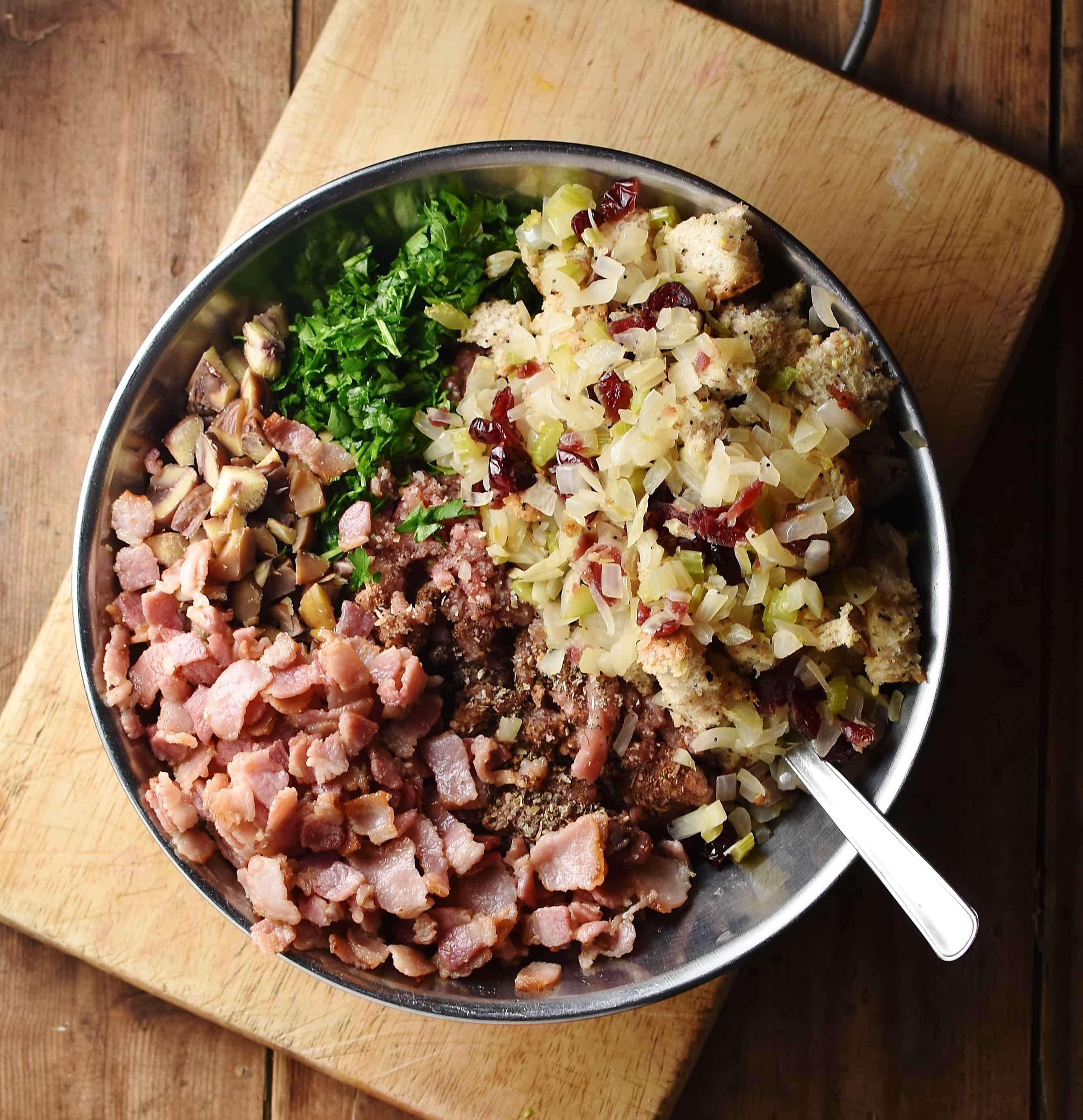 Bacon pieces, herbs, ground turkey, cranberries, chestnuts and chopped onions in large metal bowl with spoon on top of wooden board.
