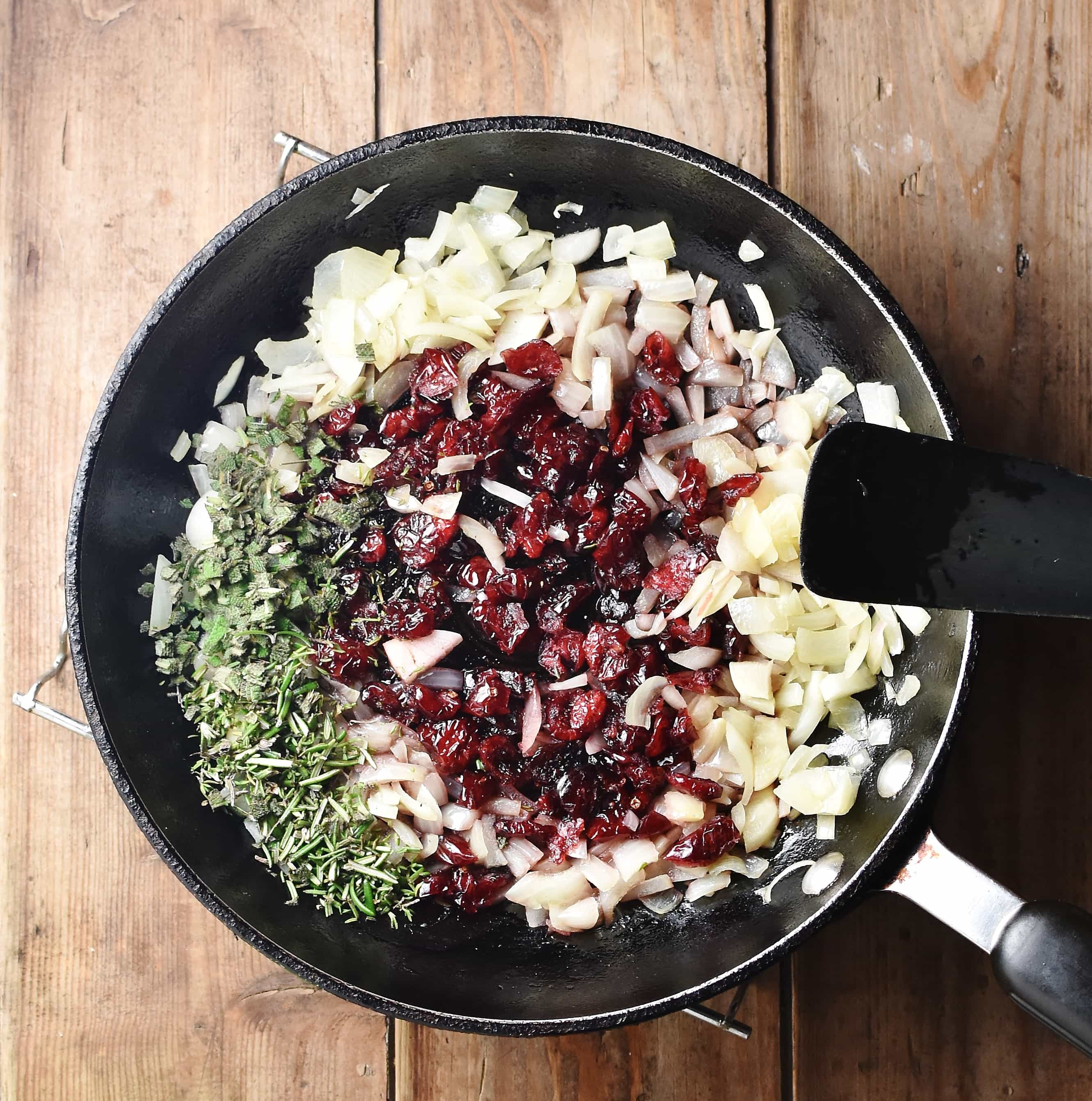 Chopped onion, cranberries and herbs in skillet with black spatula.