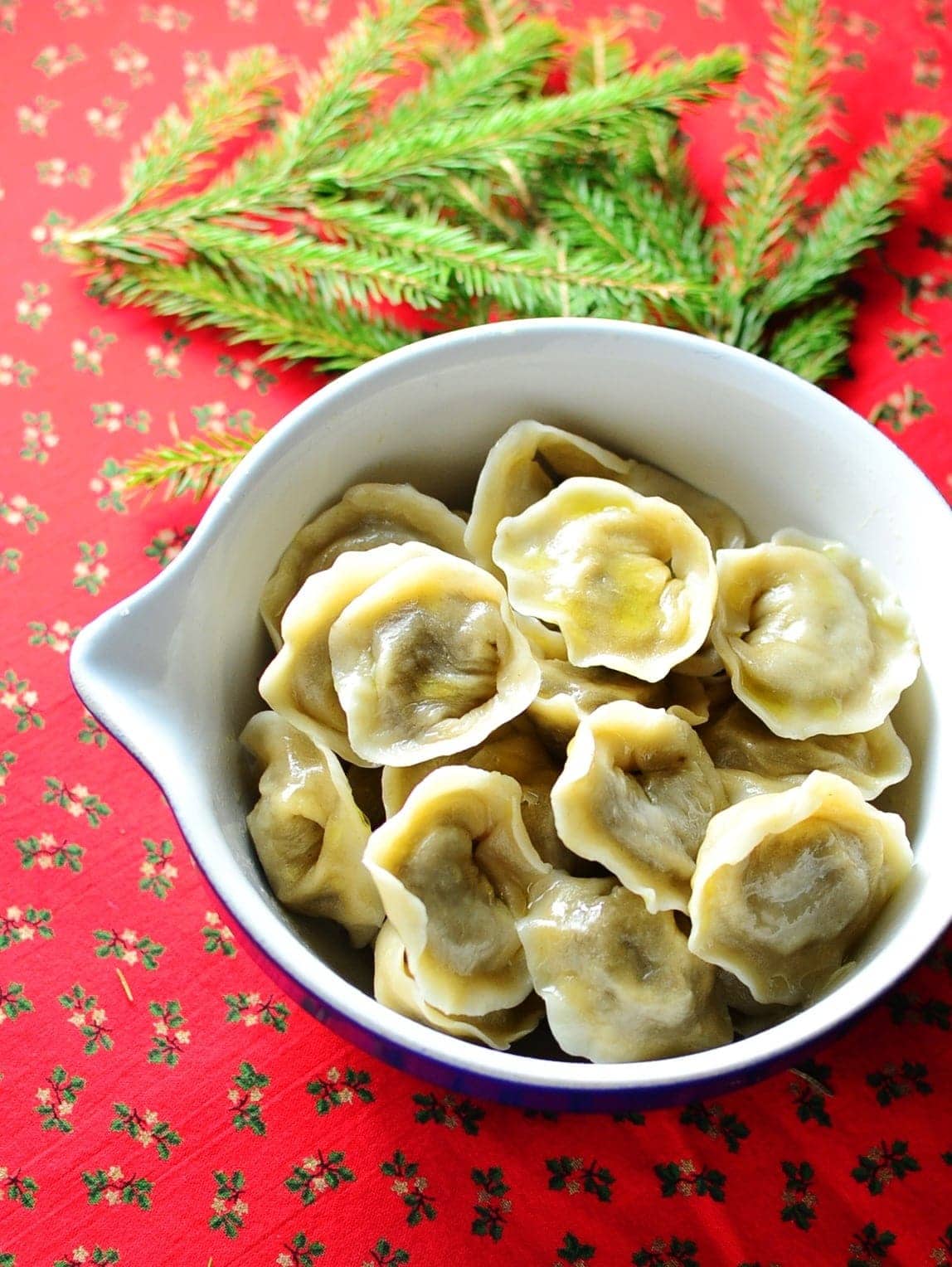 Polish Christmas Eve porcini dumplings (uszka) in white bowl with pine tree branch on red cloth with hollies pattern.