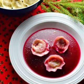 Polish borscht with Christmas uszka dumplings on white plate with uszka in bowl and pine tree branch on red cloth.