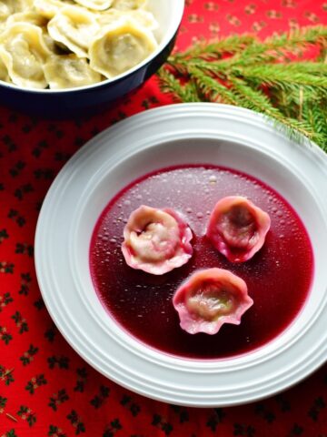 Polish borscht with Christmas uszka dumplings on white plate with uszka in bowl and pine tree branch on red cloth.