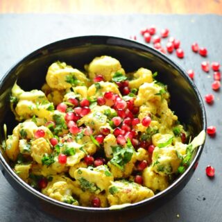 Curried cauliflower salad with yogurt dressing and pomegranate seeds in black bowl on top of grey surface.