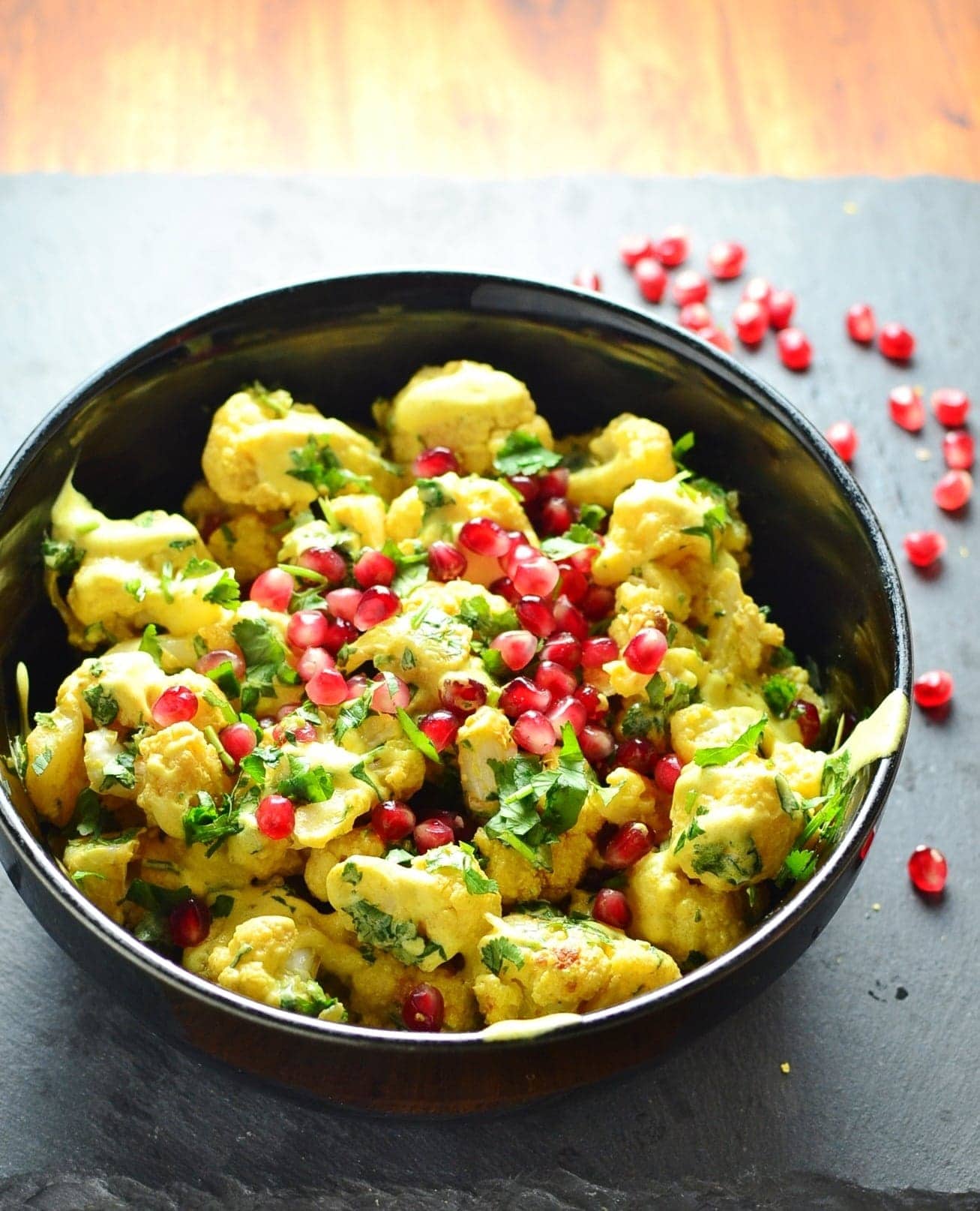 Curried cauliflower salad with yogurt dressing and pomegranate seeds in black bowl on top of grey surface.