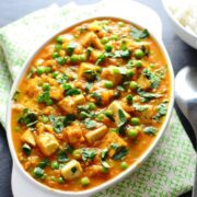 Top down view of tofu curry with peas in white oval dish on top of green-and-white cloth.