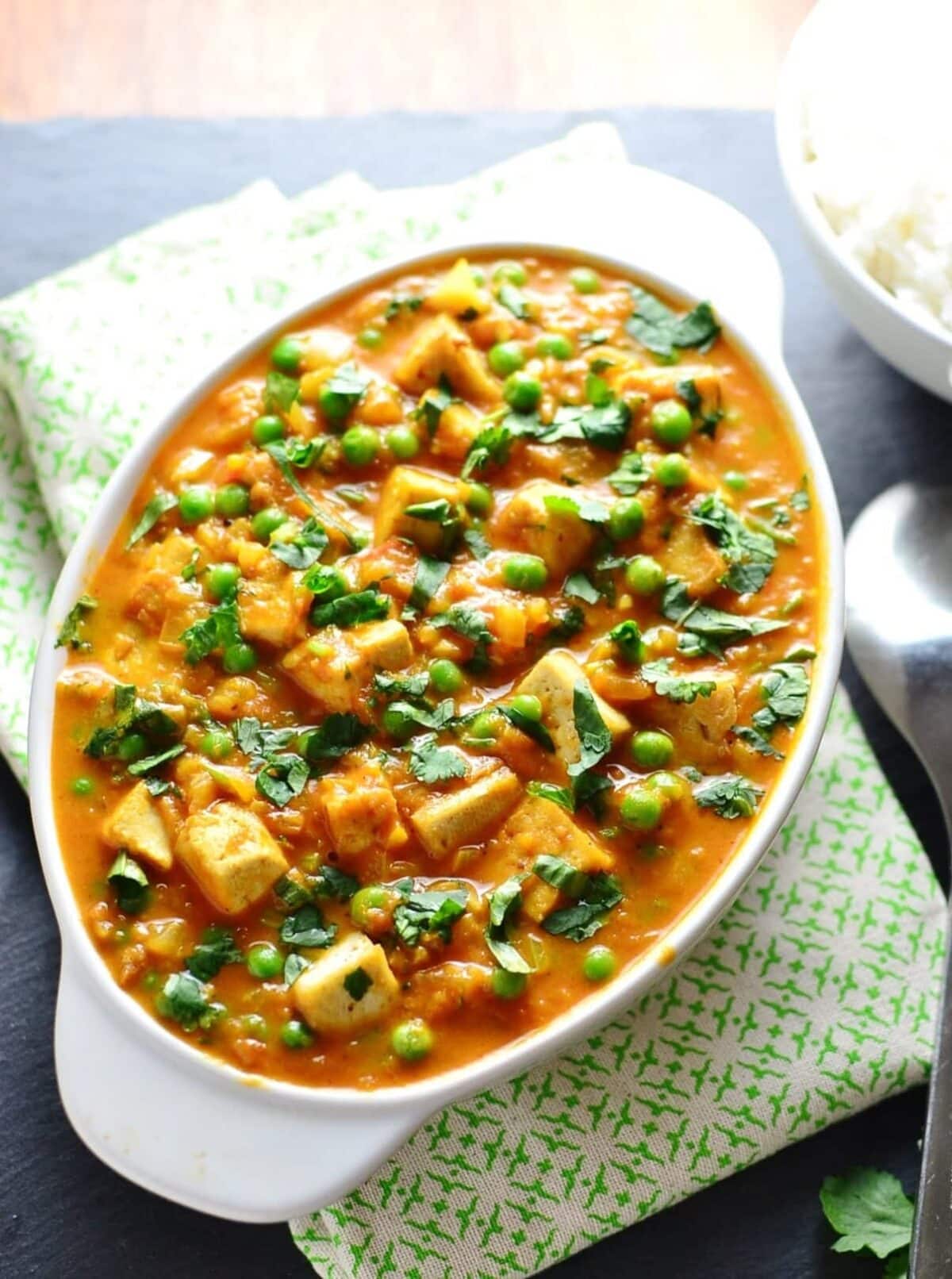 Top down view of tofu curry with peas in white oval dish on top of green-and-white cloth.