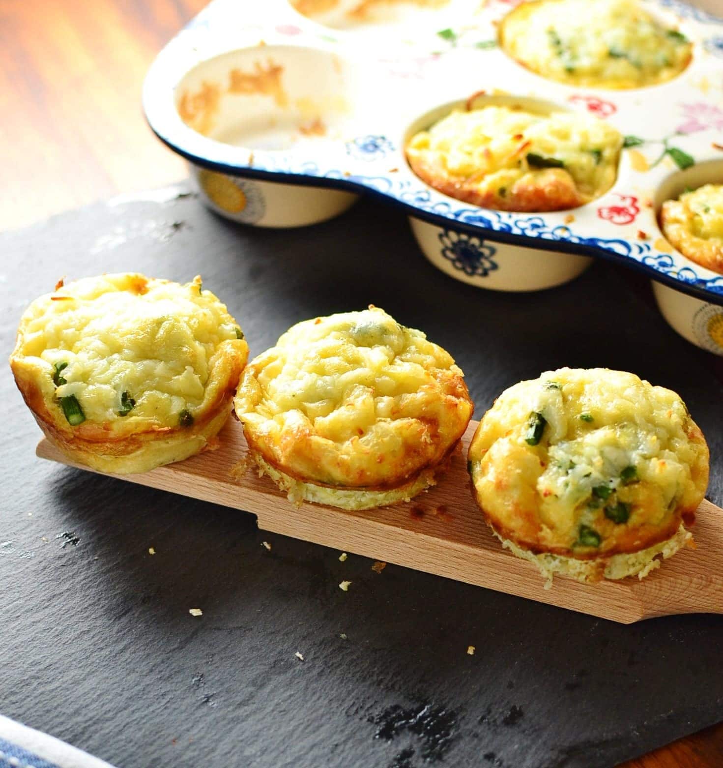 Potato asparagus frittata muffins on wooden board with ceramic muffin tin in background on slate surface.