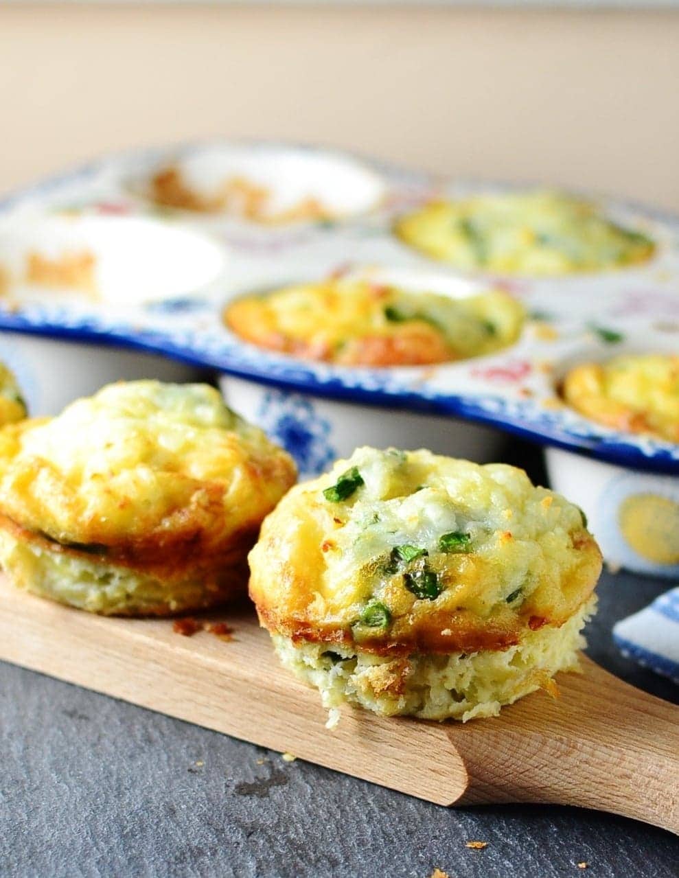 Potato asparagus frittata muffins on wooden board with ceramic muffin tin in background on dark grey surface.