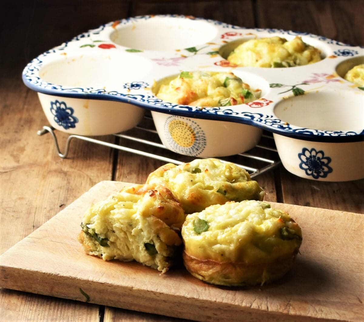 Side view of egg asparagus muffins on top of wooden board, with white ceramic muffin pan with blue flowery pattern in background.