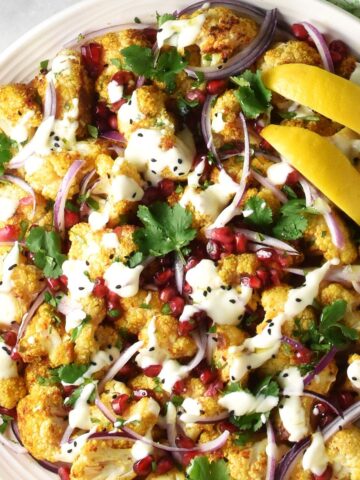 Top down close-up view of curried cauliflower salad with creamy dressing and lemon wedges on large plate.