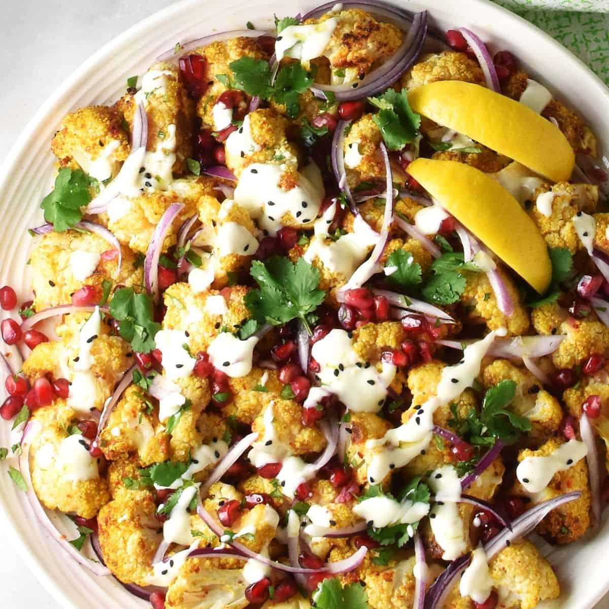 Top down close-up view of curried cauliflower salad with creamy dressing and lemon wedges on large plate.