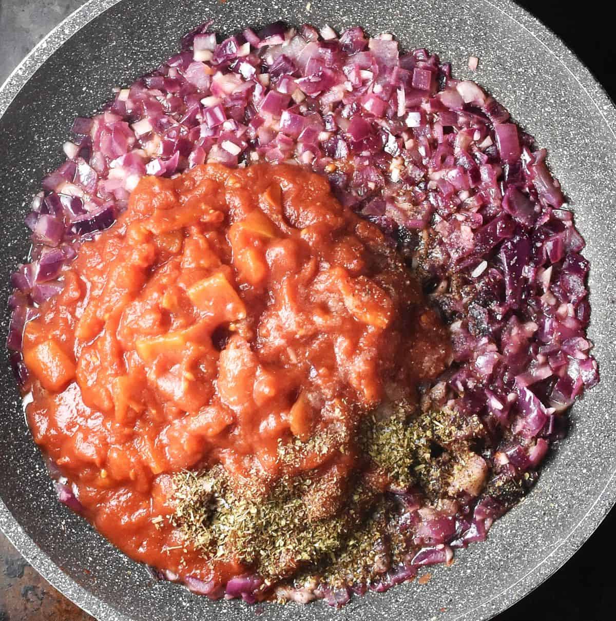 Sauteed red onion with canned tomatoes and dried herbs in large pan.