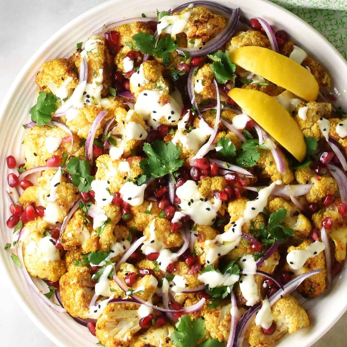 Top down view of roasted curried cauliflower salad with pomegranate, herbs and creamy dressing on large white platter.