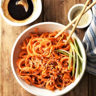 Carrot noodles stir fry in white bowl with chopsticks, seeds and glaze in background.