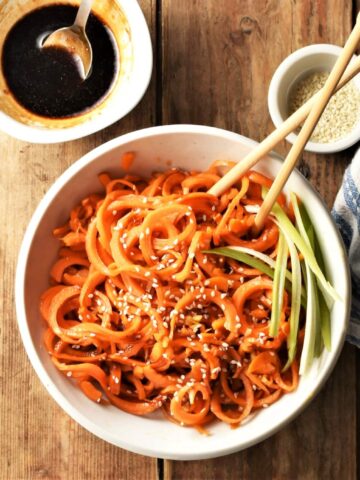 Carrot noodles stir fry in white bowl with chopsticks, seeds and glaze in background.