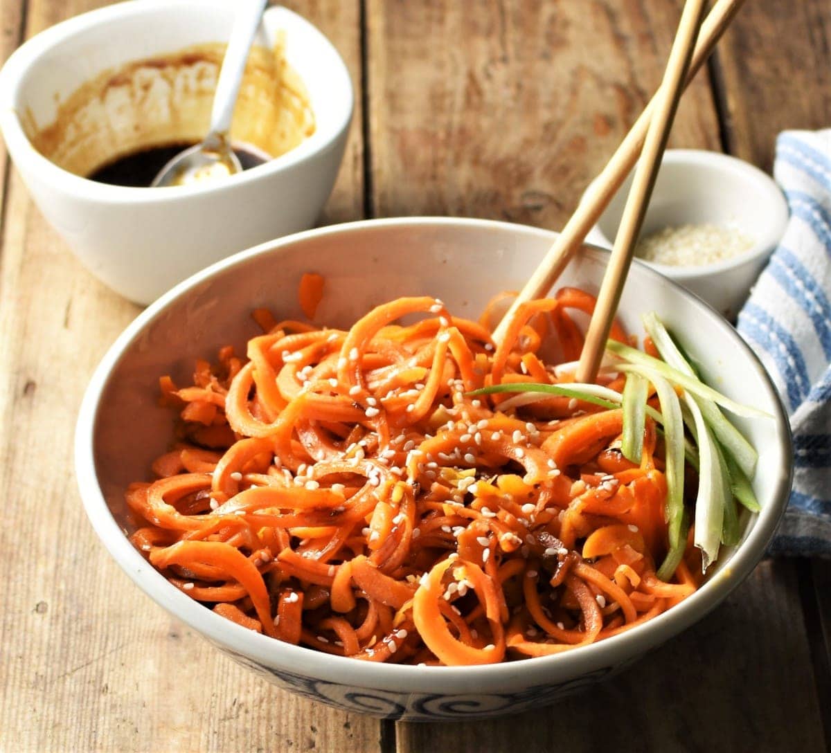 Side view of stir fried spiralized carrot in bowl with chopsticks and glaze in background.