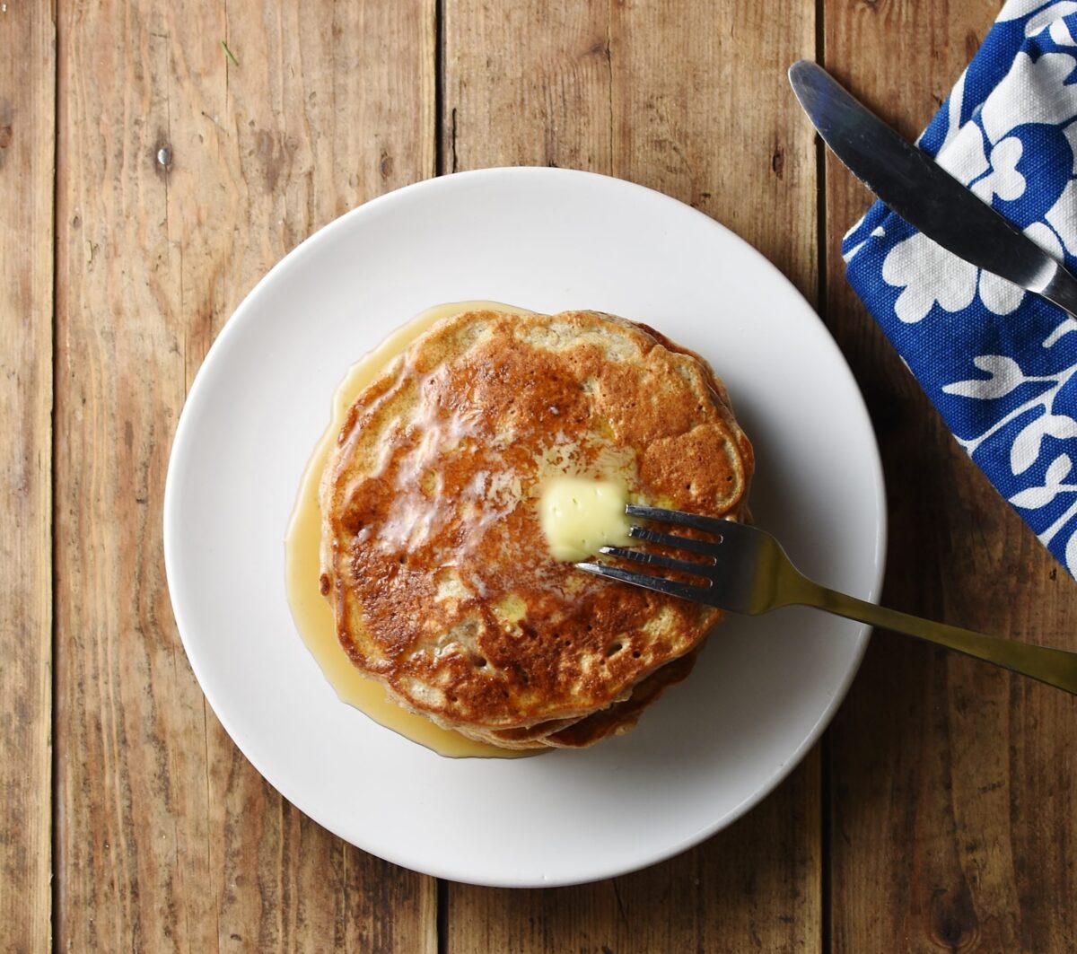 Top down view of pancakes with fork on white plate, and blue patterned cloth with knife in top right corner.