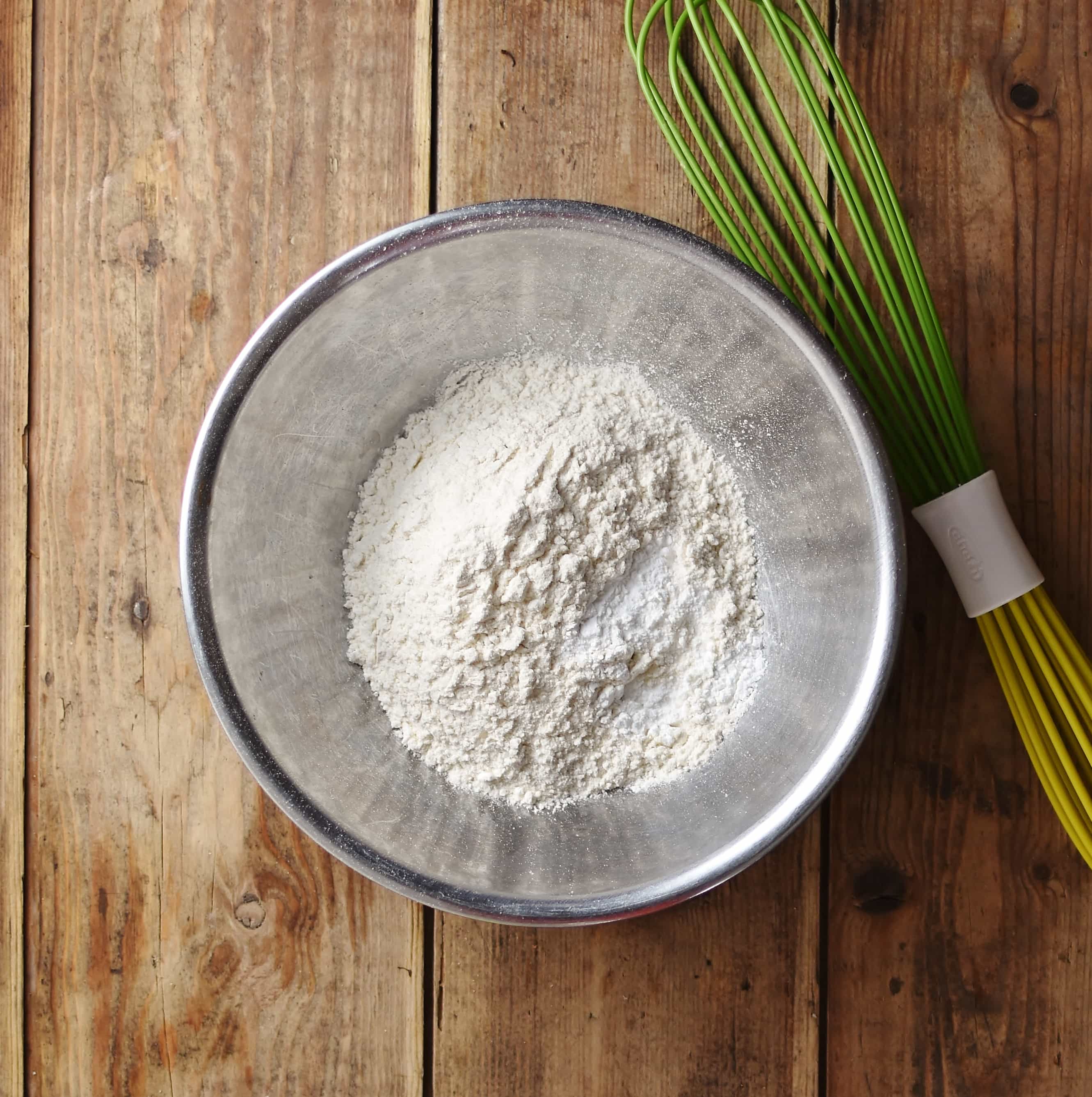 Flour inside metal bowl with green whisk next to it.