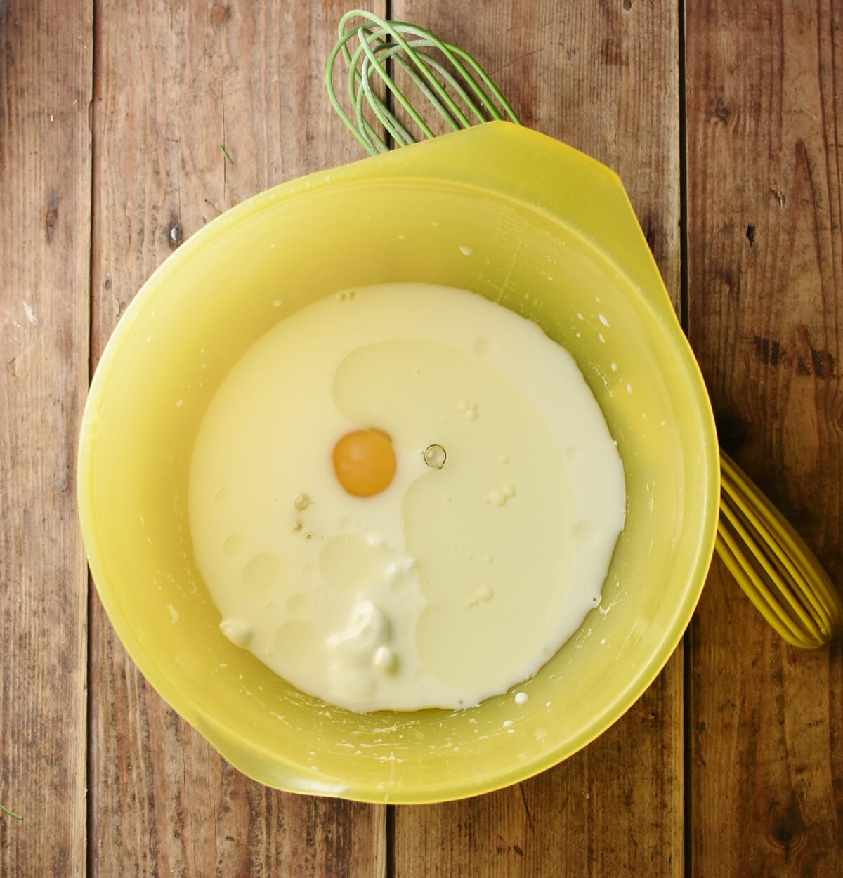 Milk and egg in large yellow bowl with green whisk to the right.