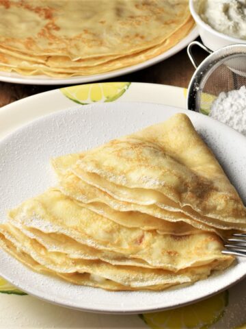 Side view of Polish crepes on plate with powdered sugar and crepes in background.