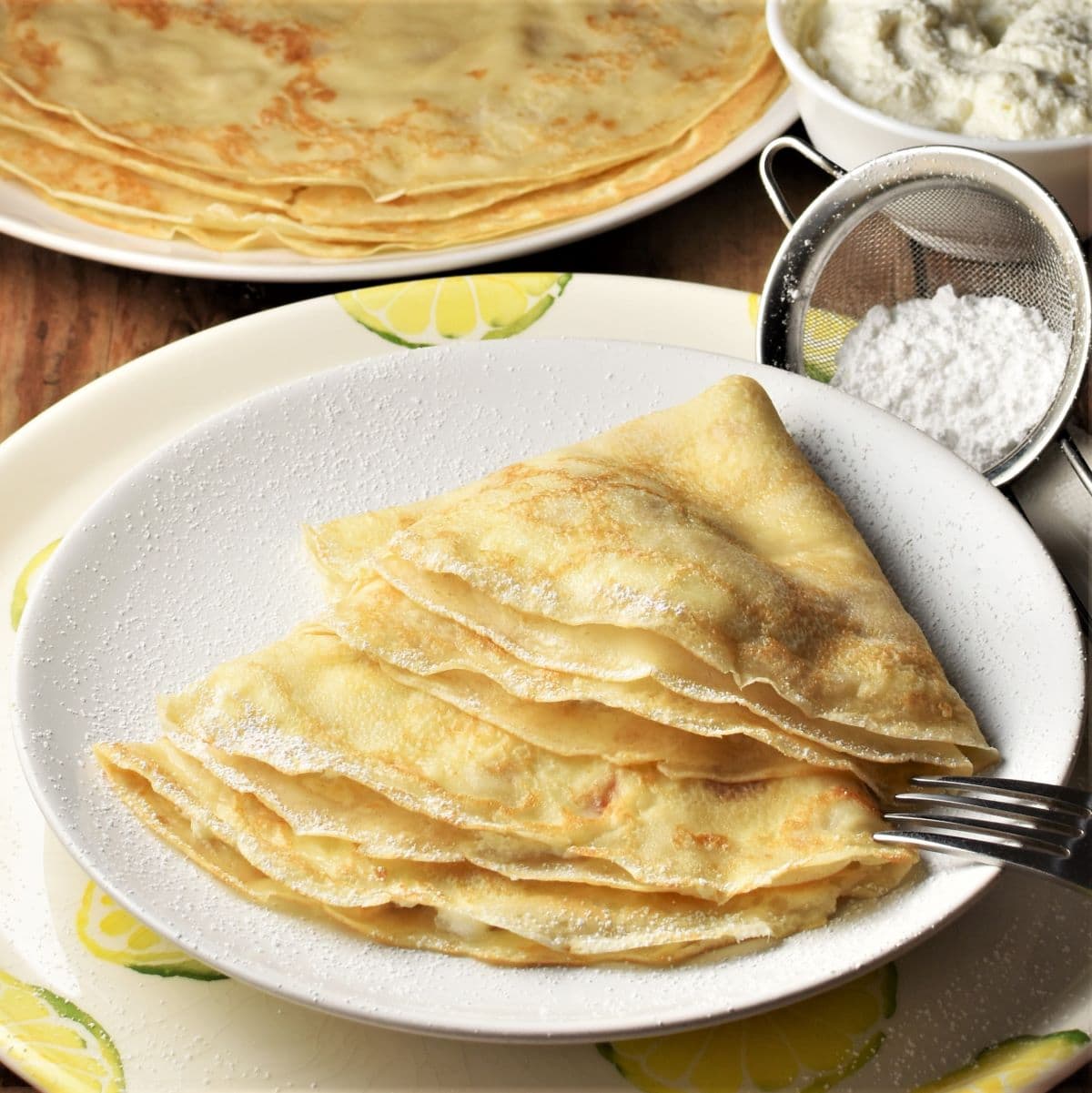 Side view of cheese crepes on plate with powdered sugar and crepes in background.