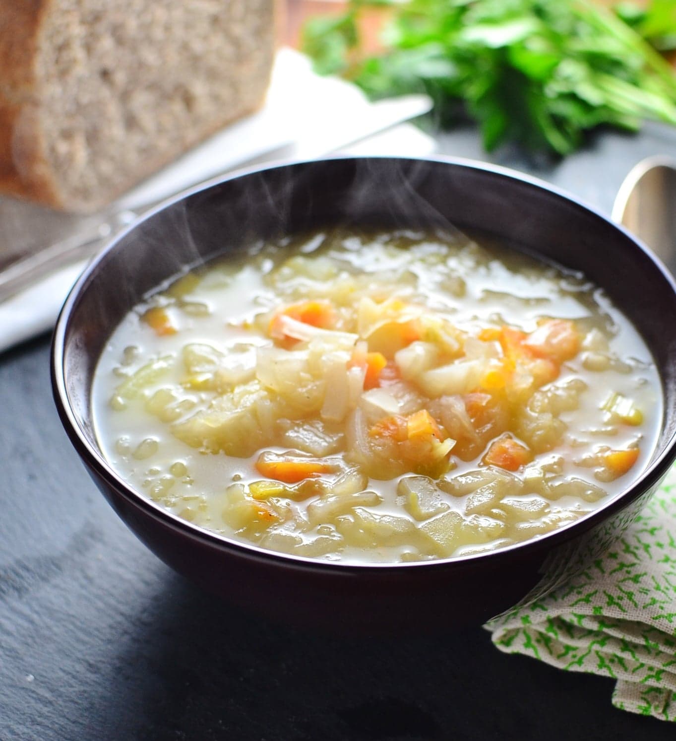 Sauerkraut vegetable soup in purple bowl with bread loaf on white cloth with knife and parsley in background.