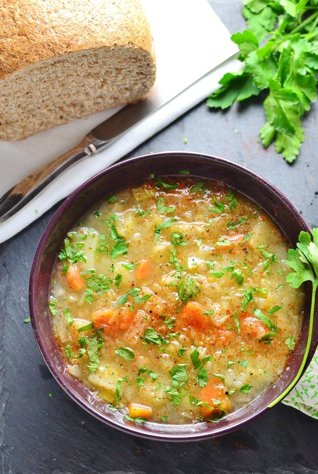 Sauerkraut vegetable kapusniak soup in purple bowl with bread loaf on white cloth with knife and parsley.