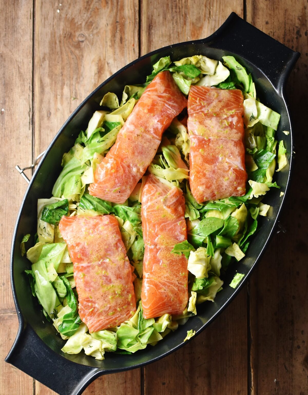 Unbaked 4 salmon pieces on bed of cabbage in black oval dish.