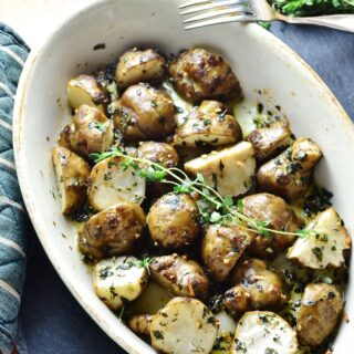 Roasted Jerusalem artichoke halves with lemon thyme in white oval dish with fork and fresh herbs in top right corner on dark grey surface.