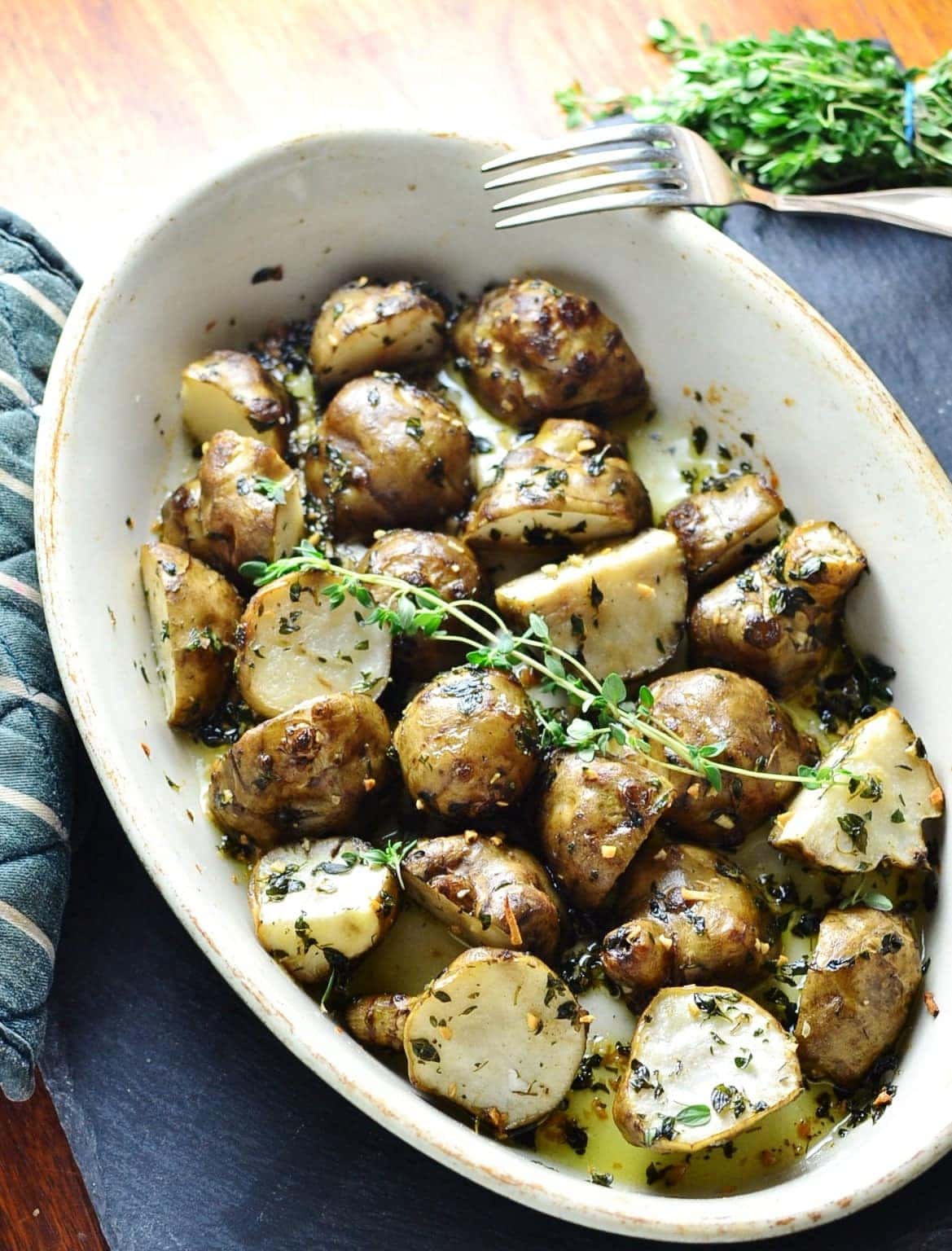 Roasted Jerusalem artichoke halves with lemon thyme in white oval dish with fork and fresh herbs in top right corner on dark grey surface. 