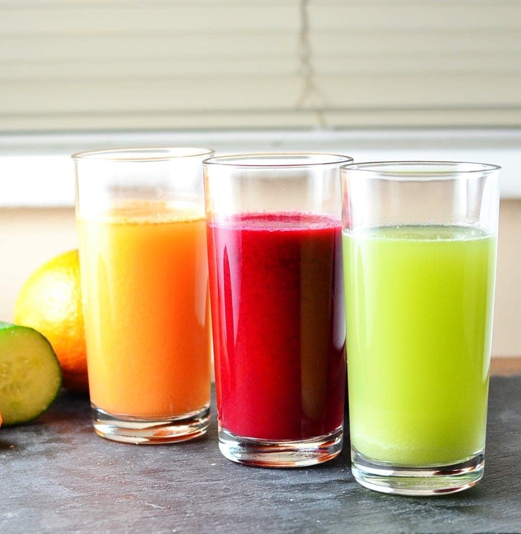 Cabbage juice in three glasses, green, red and orange, with fruit and vegetables in background on slate surface.