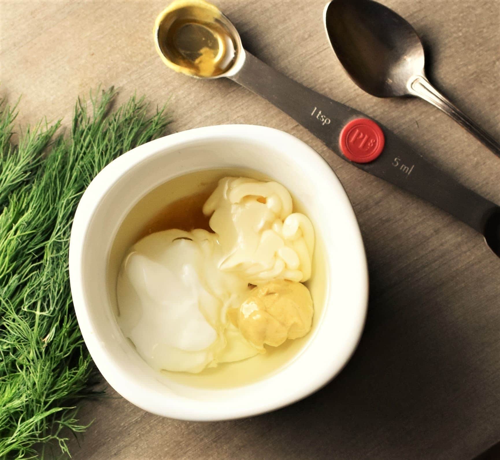 Yogurt and mustard dressing ingredients in white dish, with dill and spoons in background.