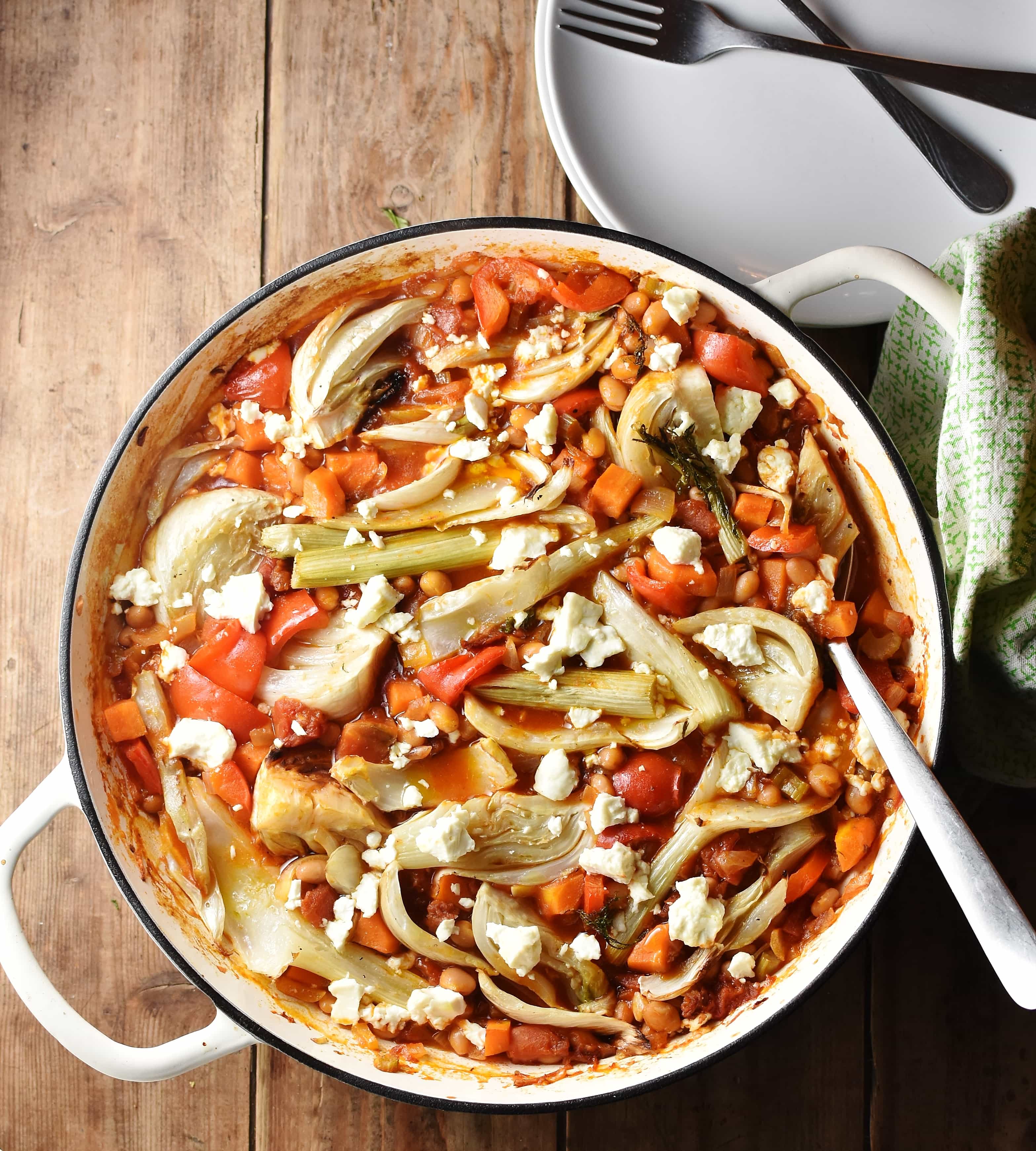 Top down view of fennel and vegetable casserole in large shallow white dish with spoon, green cloth to the right and white plate with forks in top right.