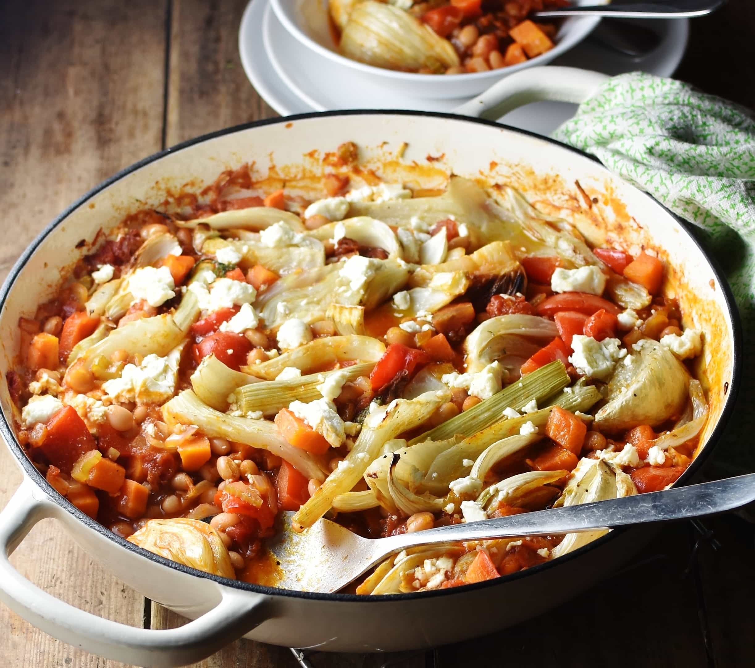 Side view of vegetable casserole with fennel pieces and beans in large shallow white dish with spoon, and white plates and casserole in bowl in background.