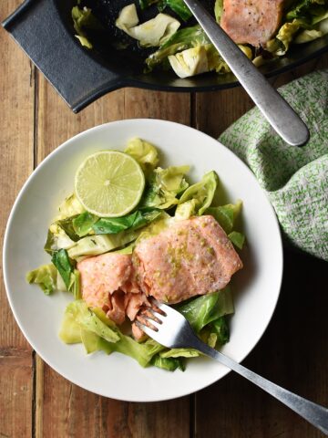 Salmon with chopped cabbage and fork in white bowl, with green cloth and black dish with salmon in background.