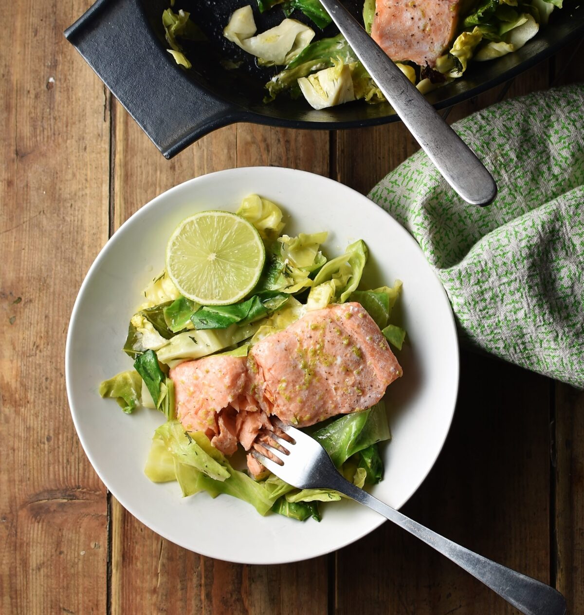 Salmon with chopped cabbage and fork in white bowl, with green cloth and black dish with salmon in background.