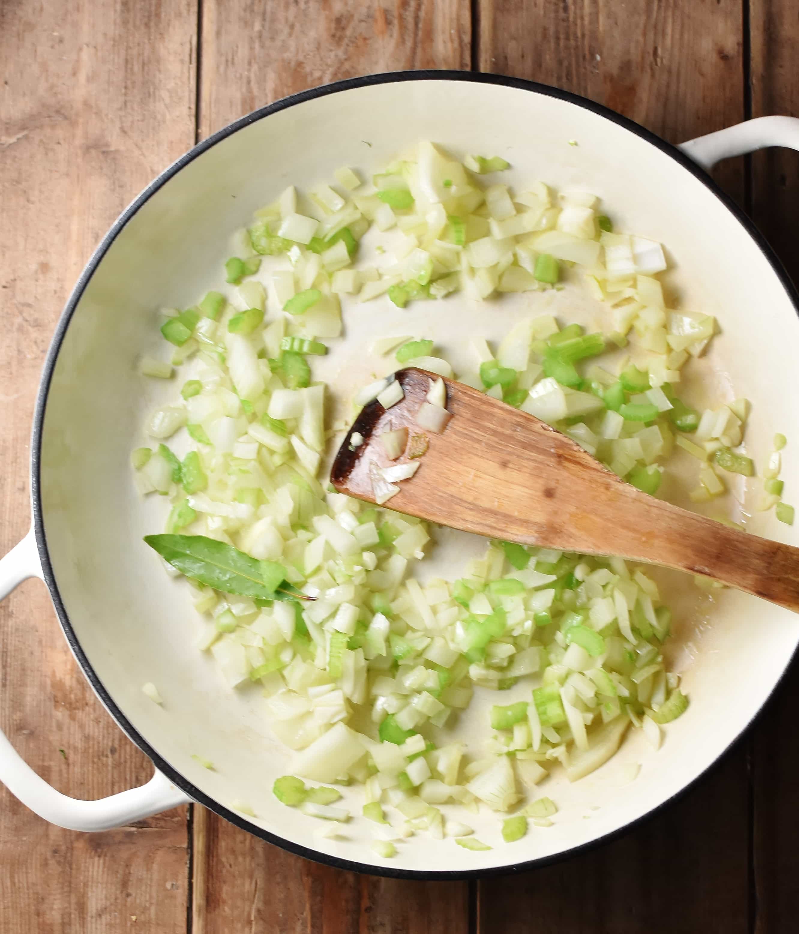 Chopped onions, celery and bay leaf in large shallow casserole with wooden spatula.