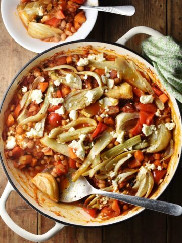 Top down view of bean, vegetable and fennel casserole with pieces of feta in large shallow white dish with spoon, green cloth in top right and casserole in small bowl at the top.