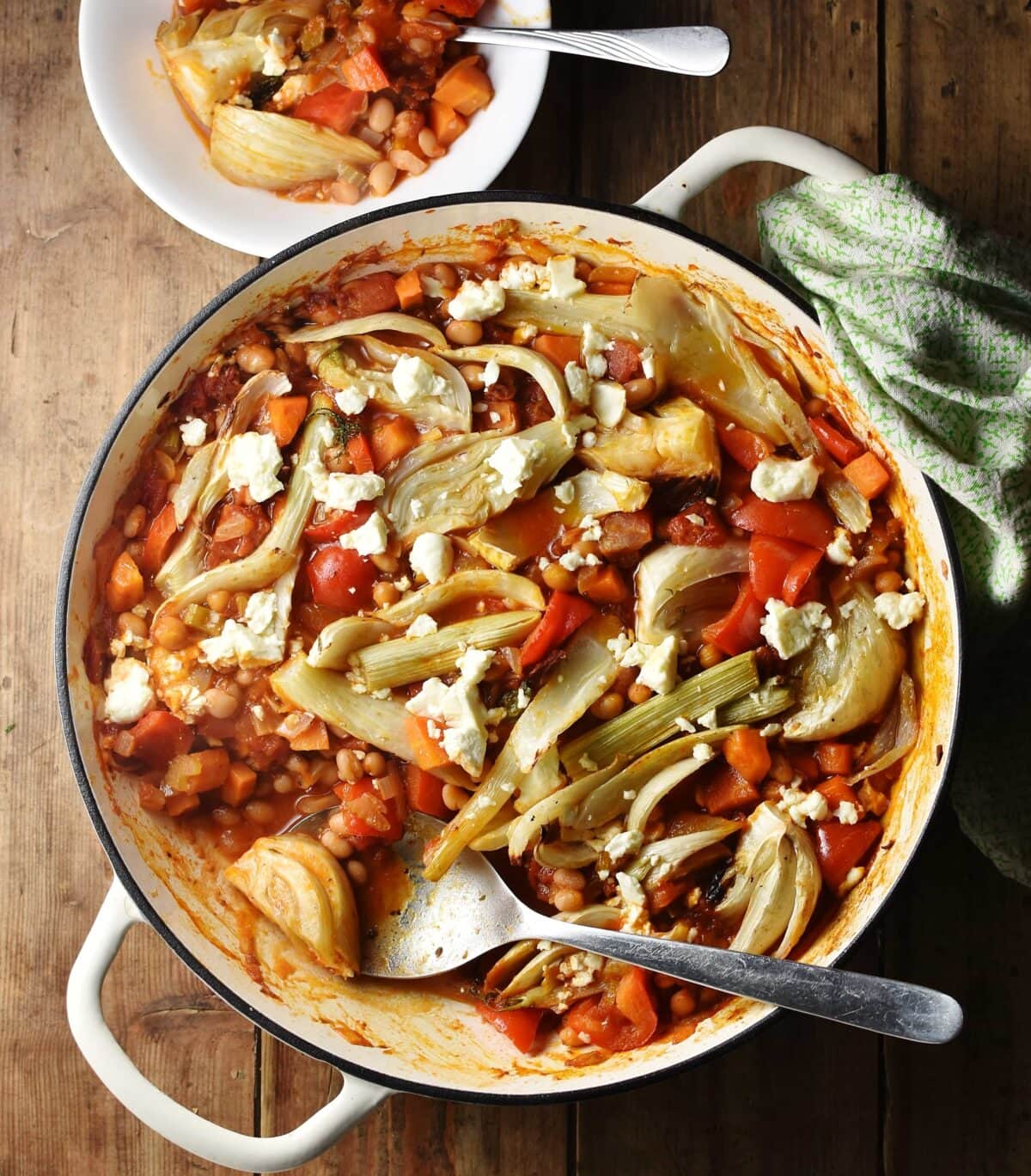 Top down view of bean, vegetable and fennel casserole with pieces of feta in large shallow white dish with spoon, green cloth in top right and casserole in small bowl at the top.