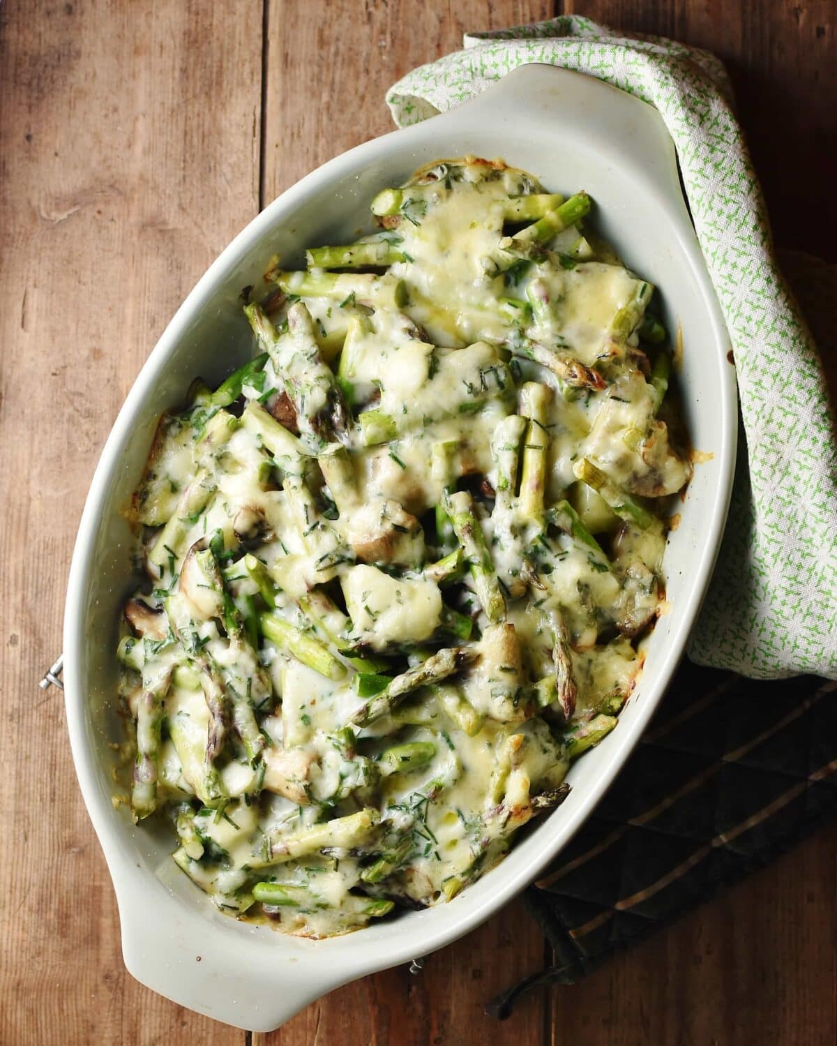 Asparagus and mushrooms with melted cheese on top in green oval dish with green cloth in top right corner.