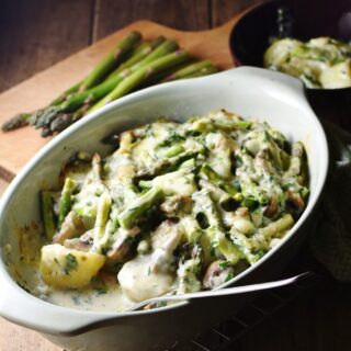 Side view of asparagus mushroom casserole with potatoes and spoon in oval dish, with asparagus tips in background.