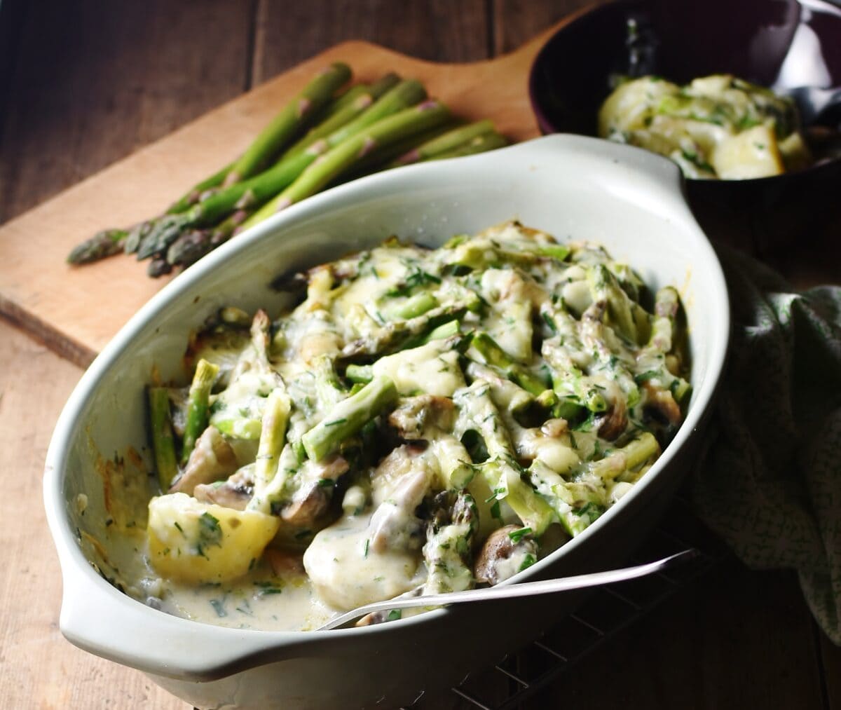 Side view of asparagus mushroom casserole with potatoes and spoon in oval dish, with asparagus tips in background.