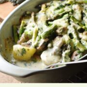 Side view of asparagus, mushroom and potato bake with creamy sauce and spoon in green oval dish, with asparagus tips in background.