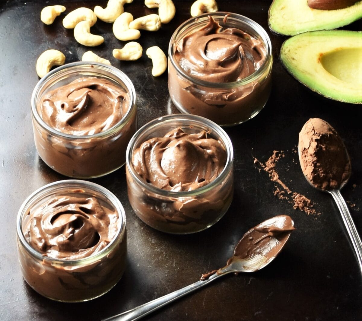 Healthy chocolate avocado mousse in 4 round pots, 2 spoons with cocoa, cashew and halved avocado.