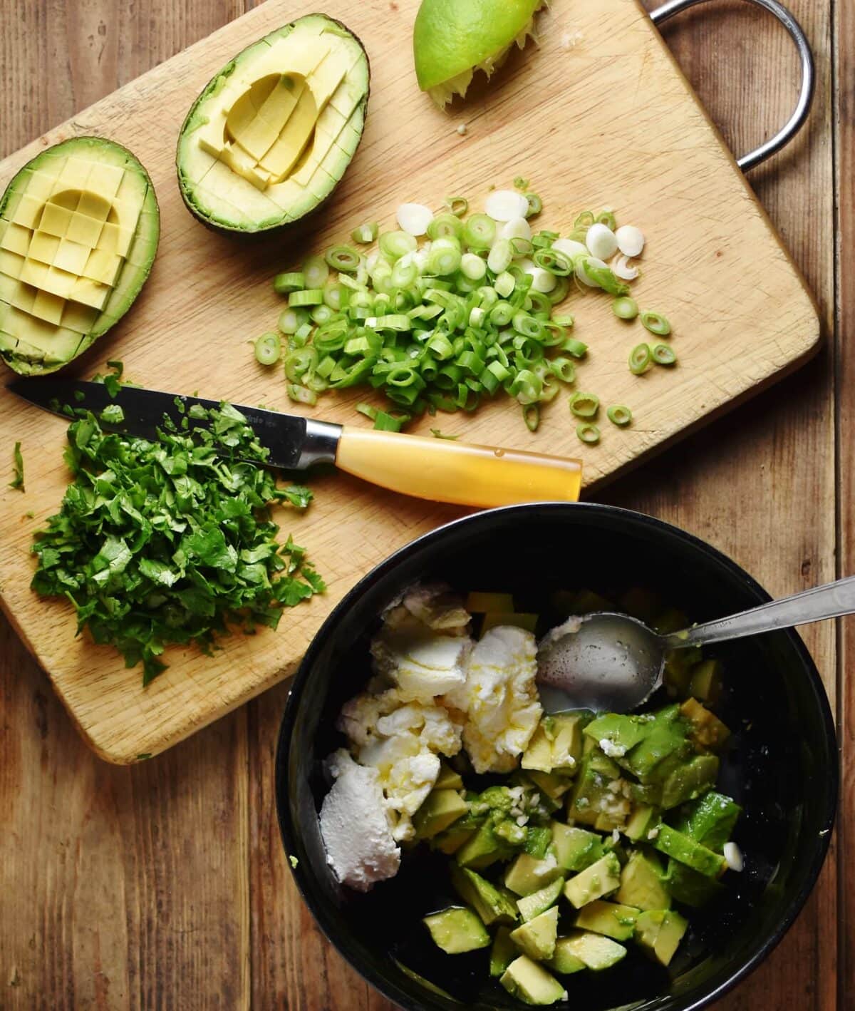 Halved avocado, onions, herbs and knife on top of cutting board and avocado, yogurt and spoon inside black bowl.