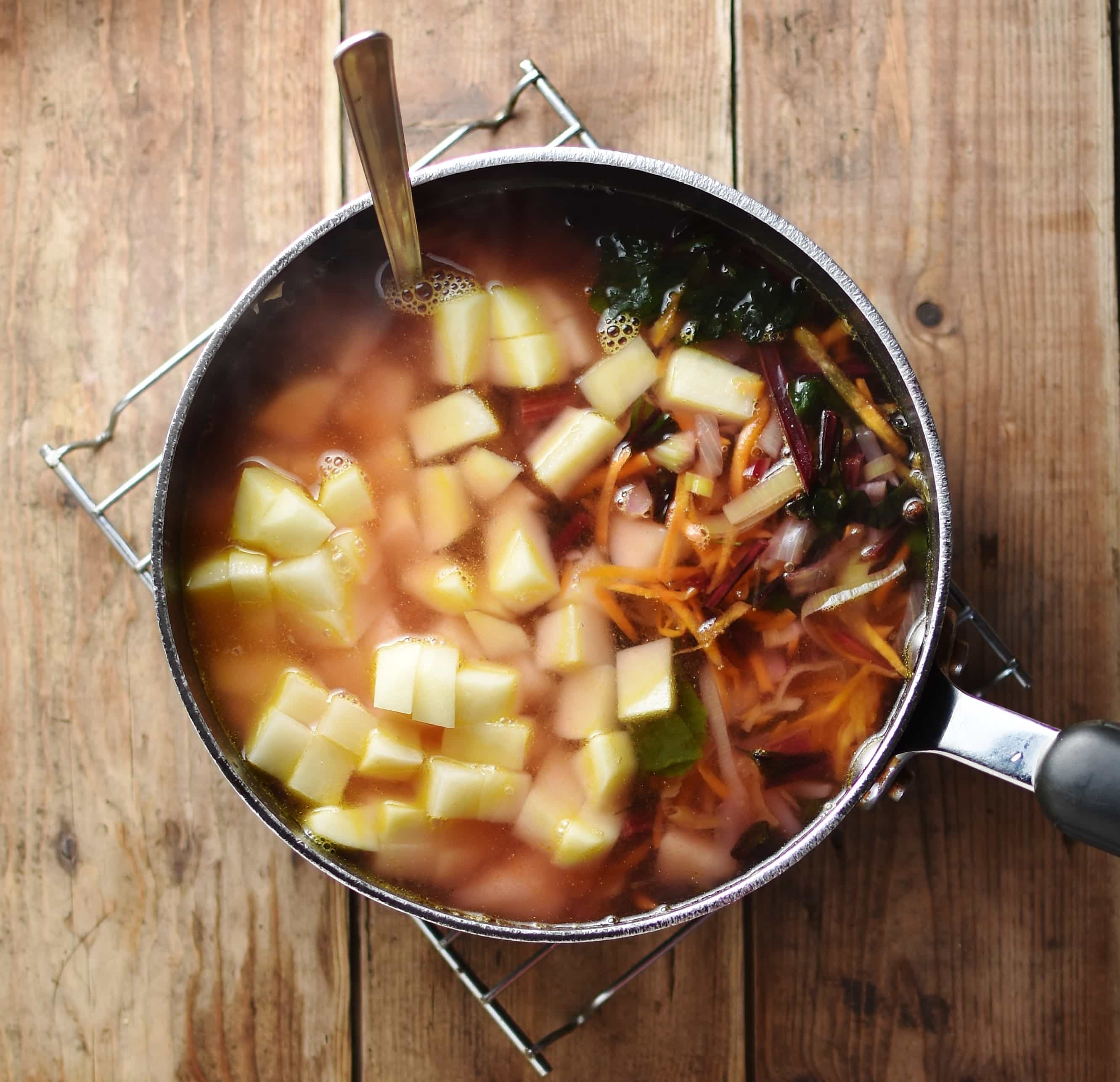 Vegetables and cubed potatoes with spoon inside soup in large pot.