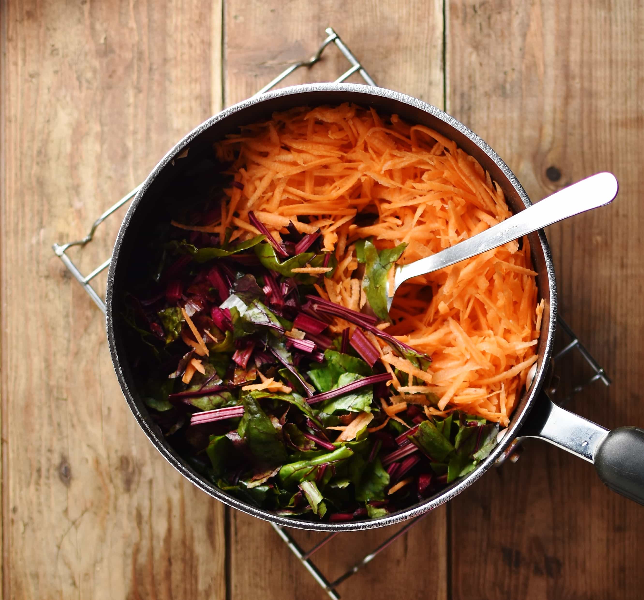 Grated carrot and chopped beet leaves with spoon inside large pot.