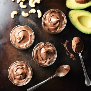 4 round chocolate mousse pots, 2 spoons with cocoa powder, cashew nuts and halved avocado.